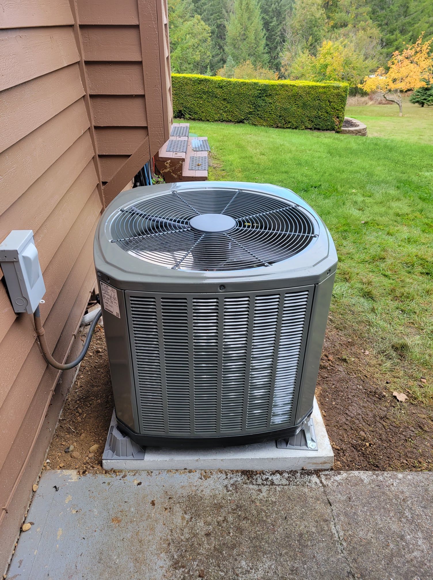 Gray air conditioning unit on a concrete pad next to a brown building. Green grass and foliage in background.