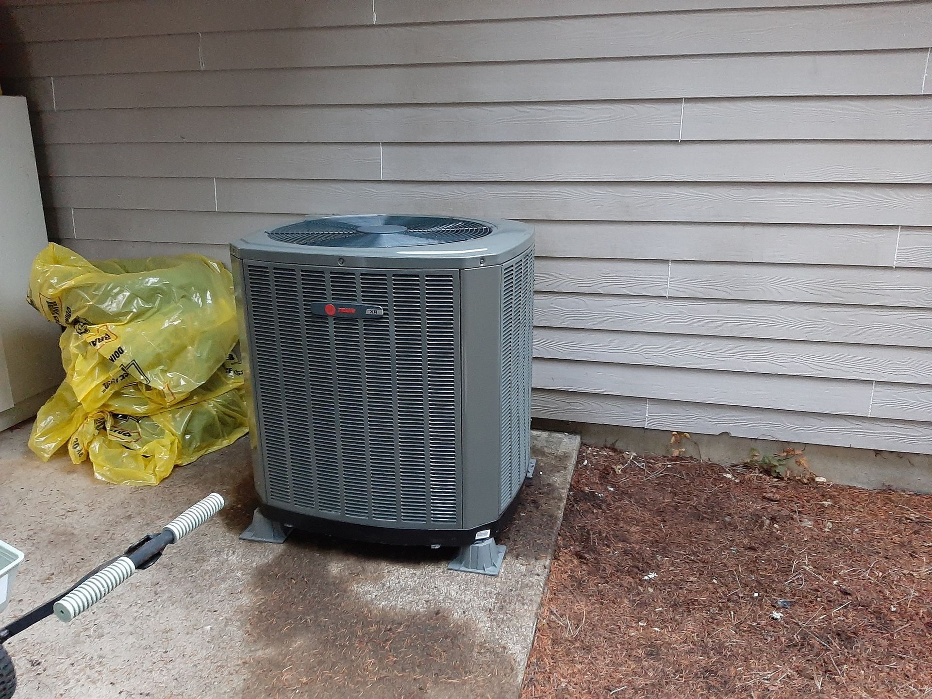 Gray air conditioning unit on concrete pad next to brown wall, with a yellow bag and hose nearby.