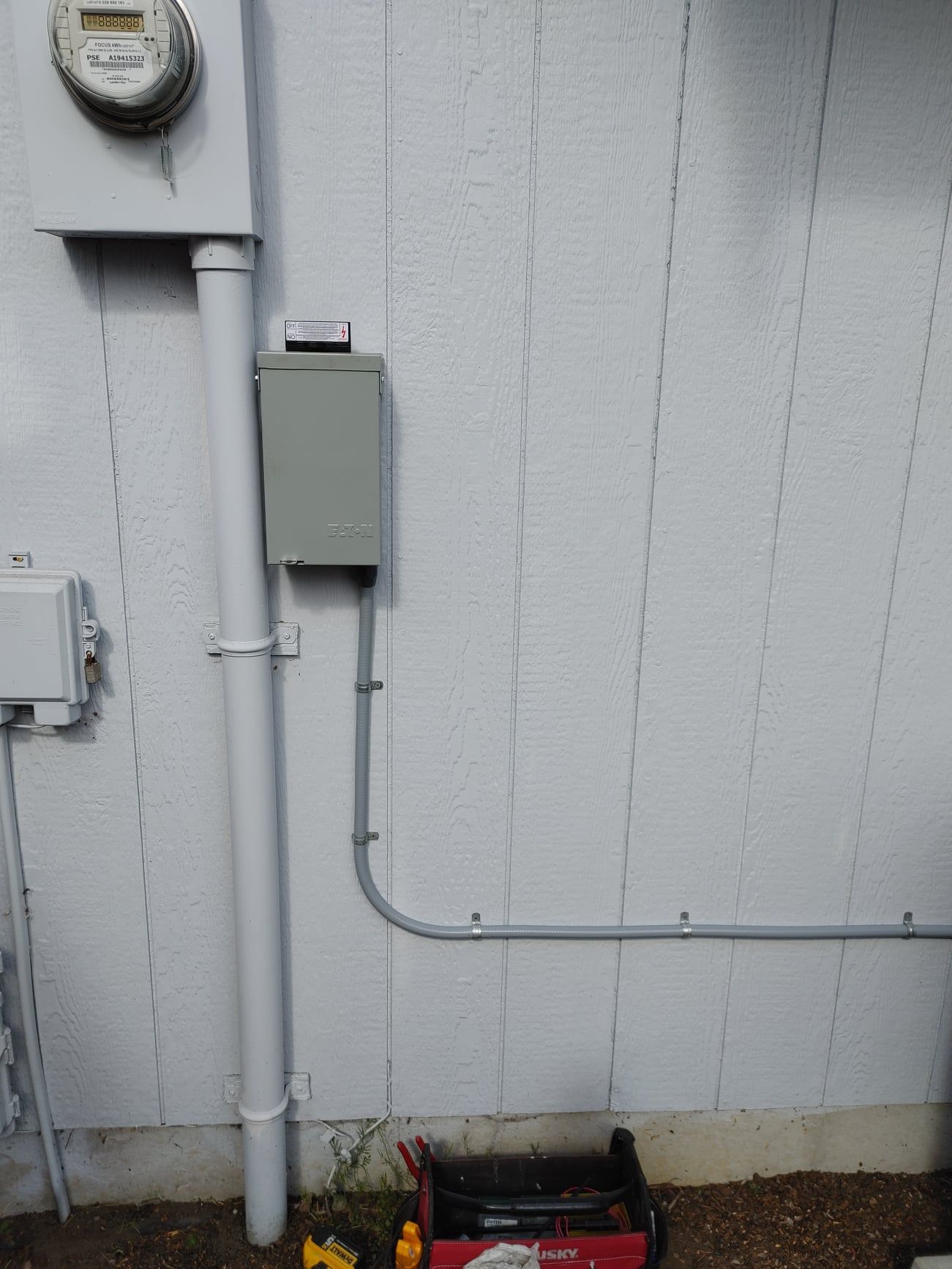 Electrical meter and service box on a gray siding wall, conduit running down and to the right, tool box below.