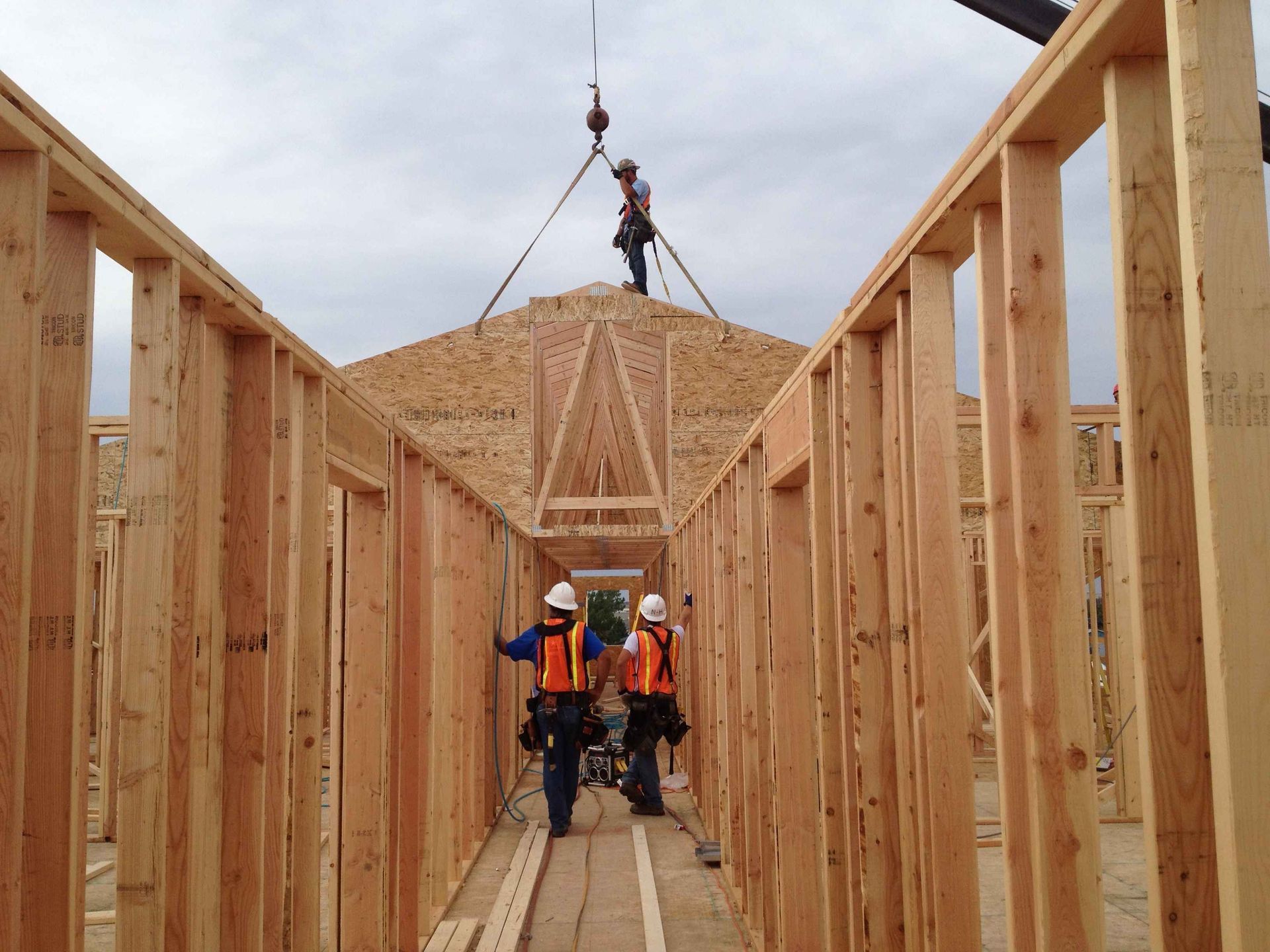 Construction workers in orange vests inside a wooden house frame, lifting materials with a crane.