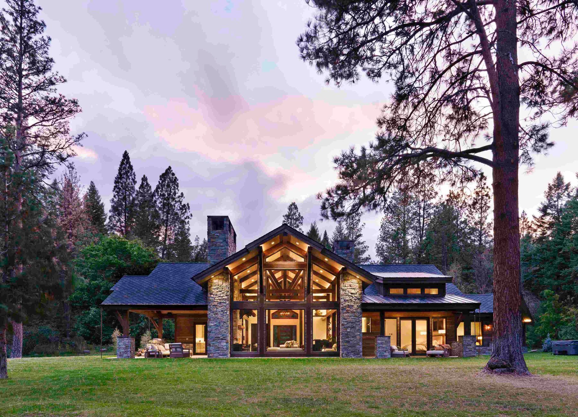 Modern cabin with large windows glowing at dusk, surrounded by tall trees and a grassy yard.
