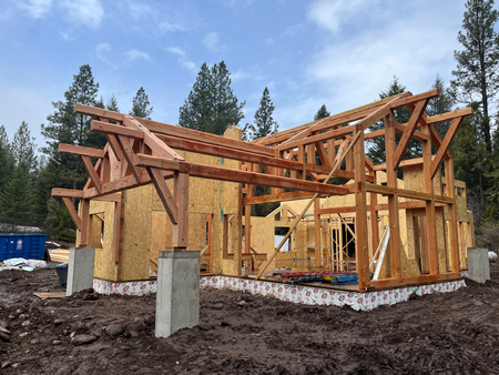 Wood-framed house under construction with exposed trusses and plywood walls on a muddy site