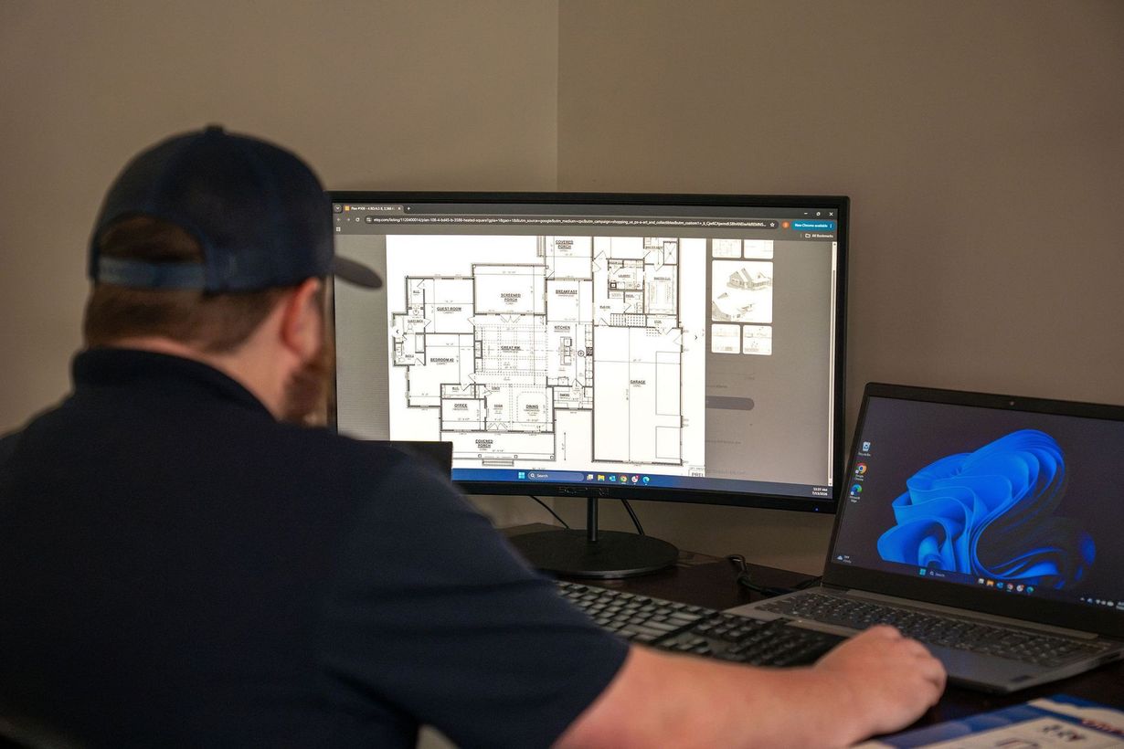 Person at a desk reviewing floor plans on a monitor and a blue 3D model on a laptop.