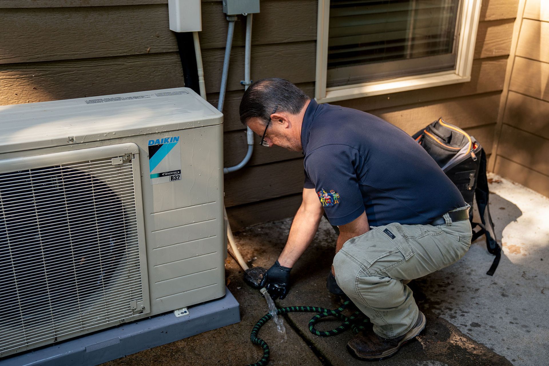 Technician kneeling beside an outdoor air conditioner, inspecting a hose and connections by a house wall