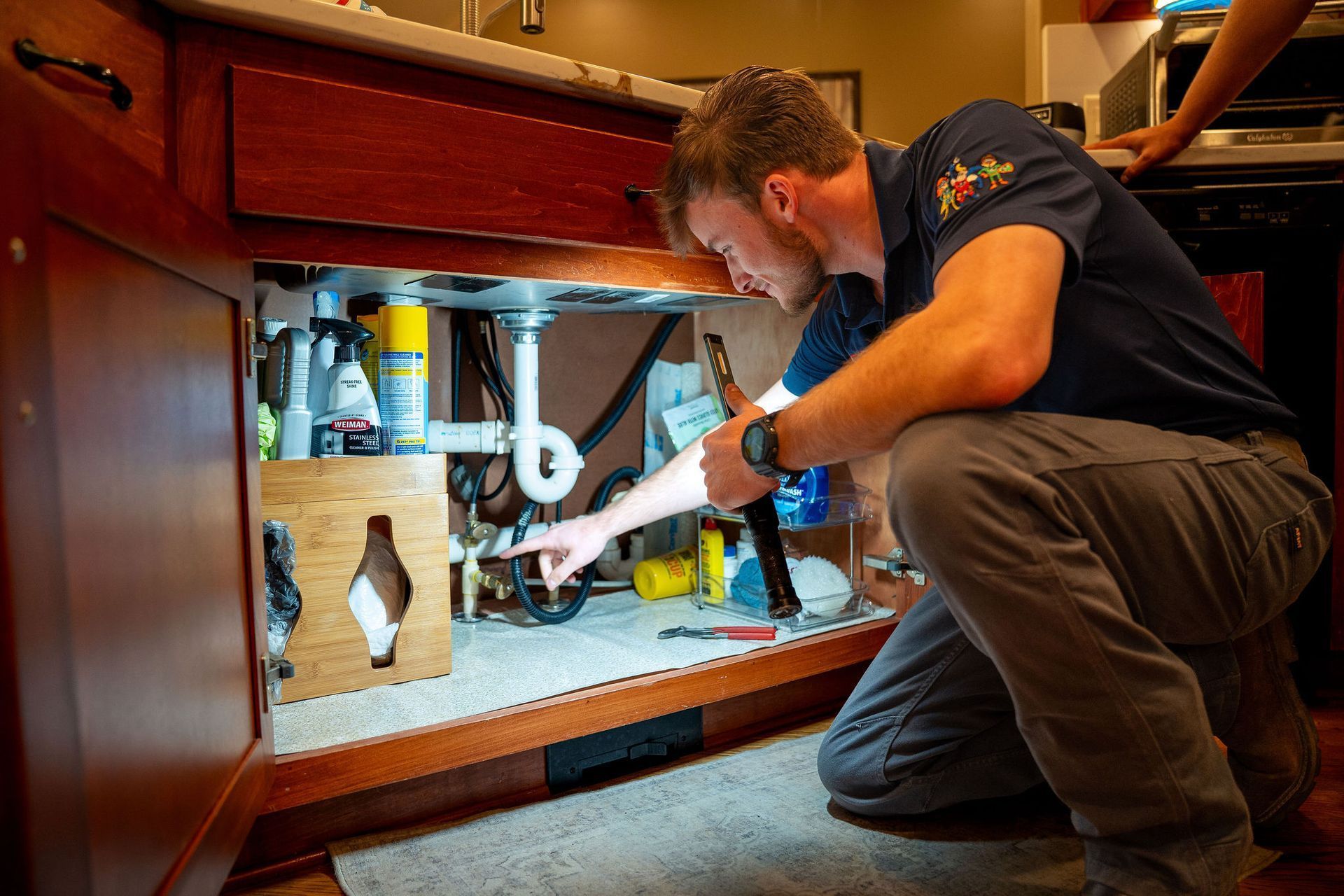 Technician crouches under a sink, inspecting plumbing tools and pipes in a cabinet.