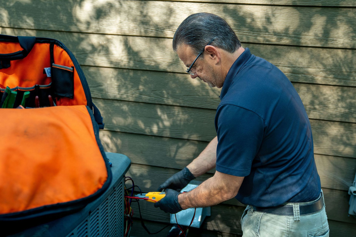 Man in blue shirt uses a tool outdoors beside an orange bag and wooden fence.