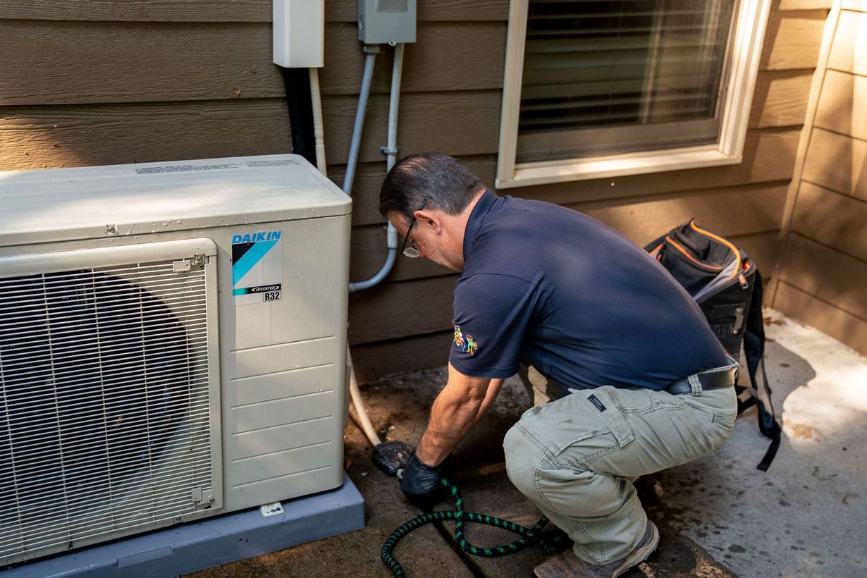 Technician kneels beside an outdoor air conditioner, inspecting hoses and tools on a patio.