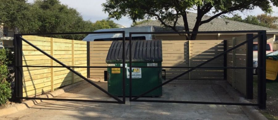 A green dumpster is behind a gate in front of a house.