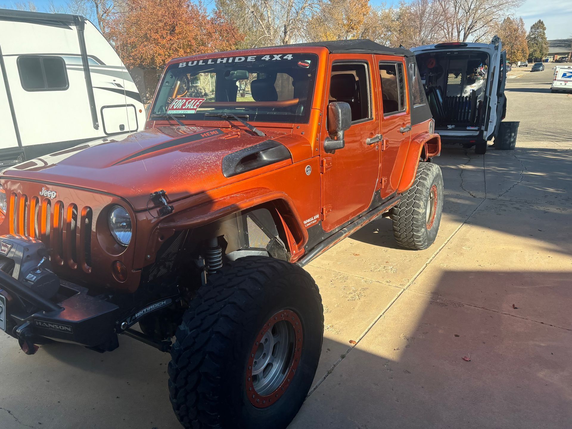 Orange Jeep Wrangler with large tires parked on pavement next to a white trailer and black vehicle.