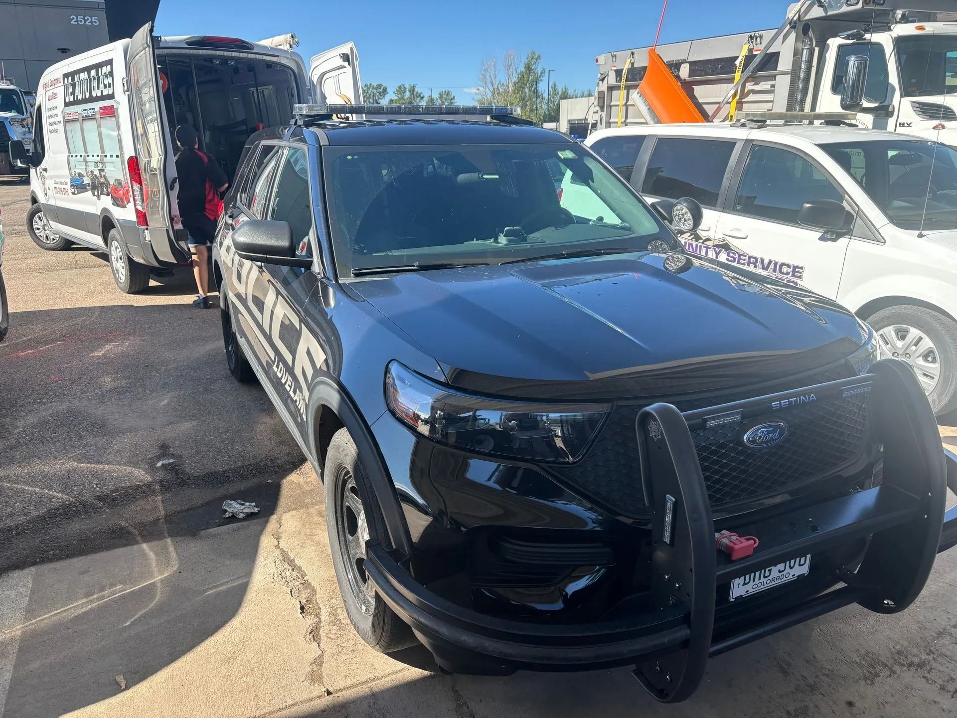 Black police SUV with push bar parked near a white van and a silver van with equipment.
