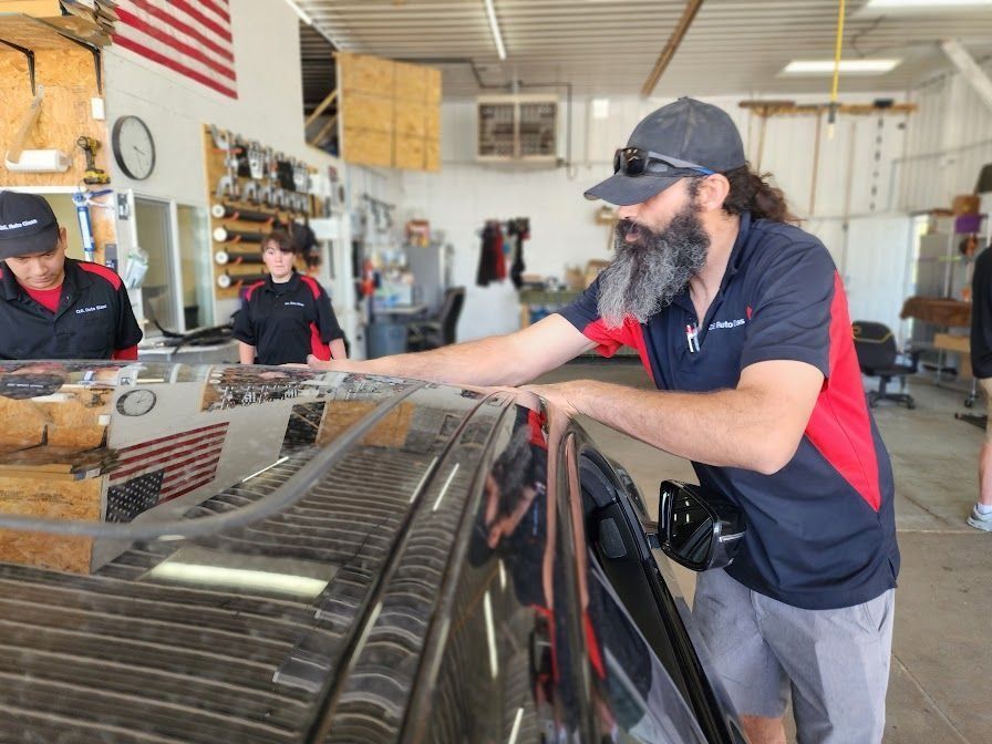 Man with beard inspecting car roof; two people in the background, shop setting.