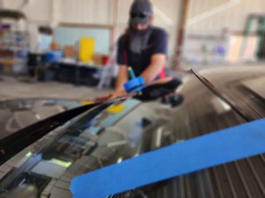 Person working on a car windshield. They are kneeling, wearing a hat, gloves, and working inside a garage.