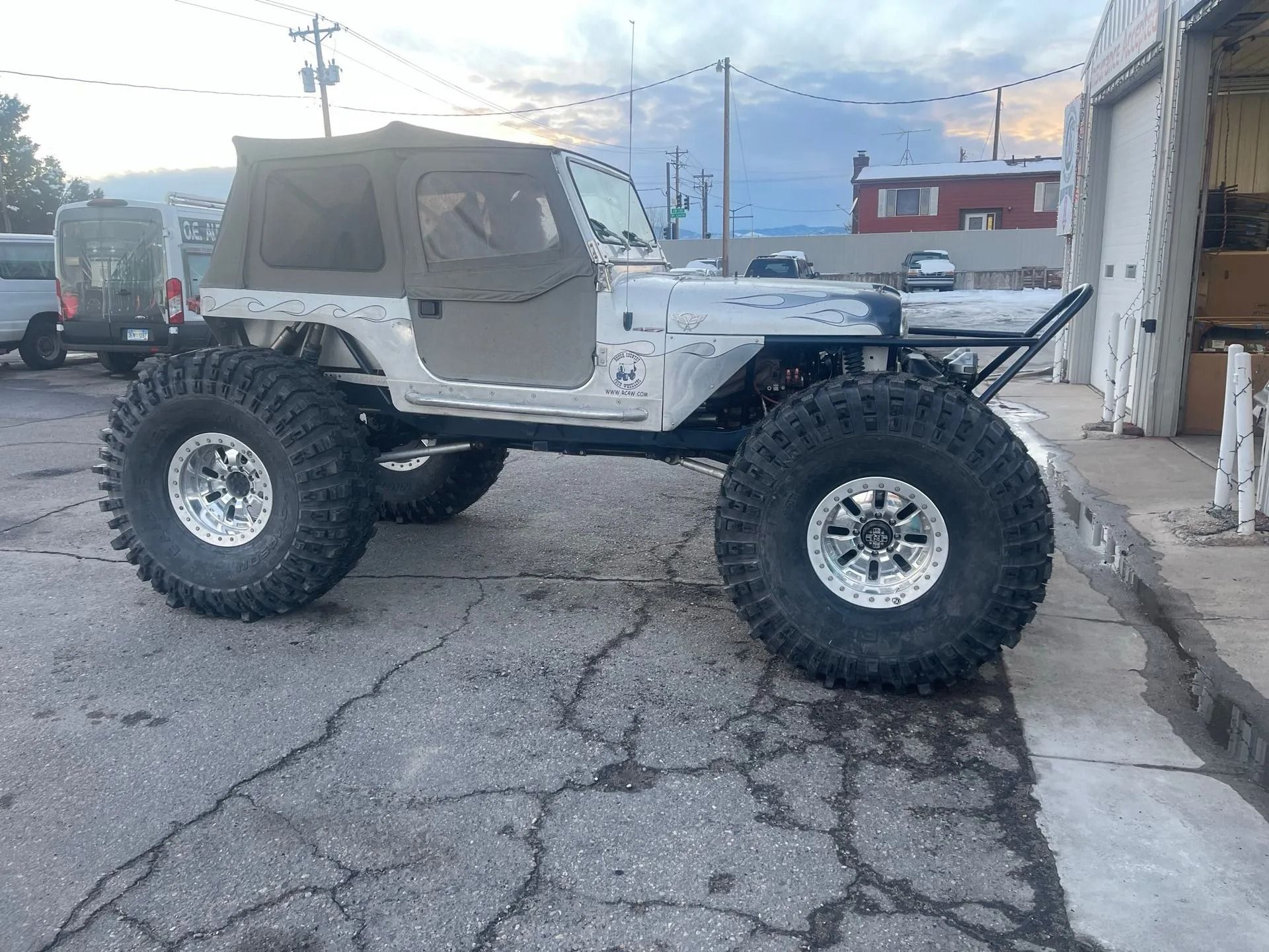 Modified silver Jeep with large tires and a black soft top parked outside a building.