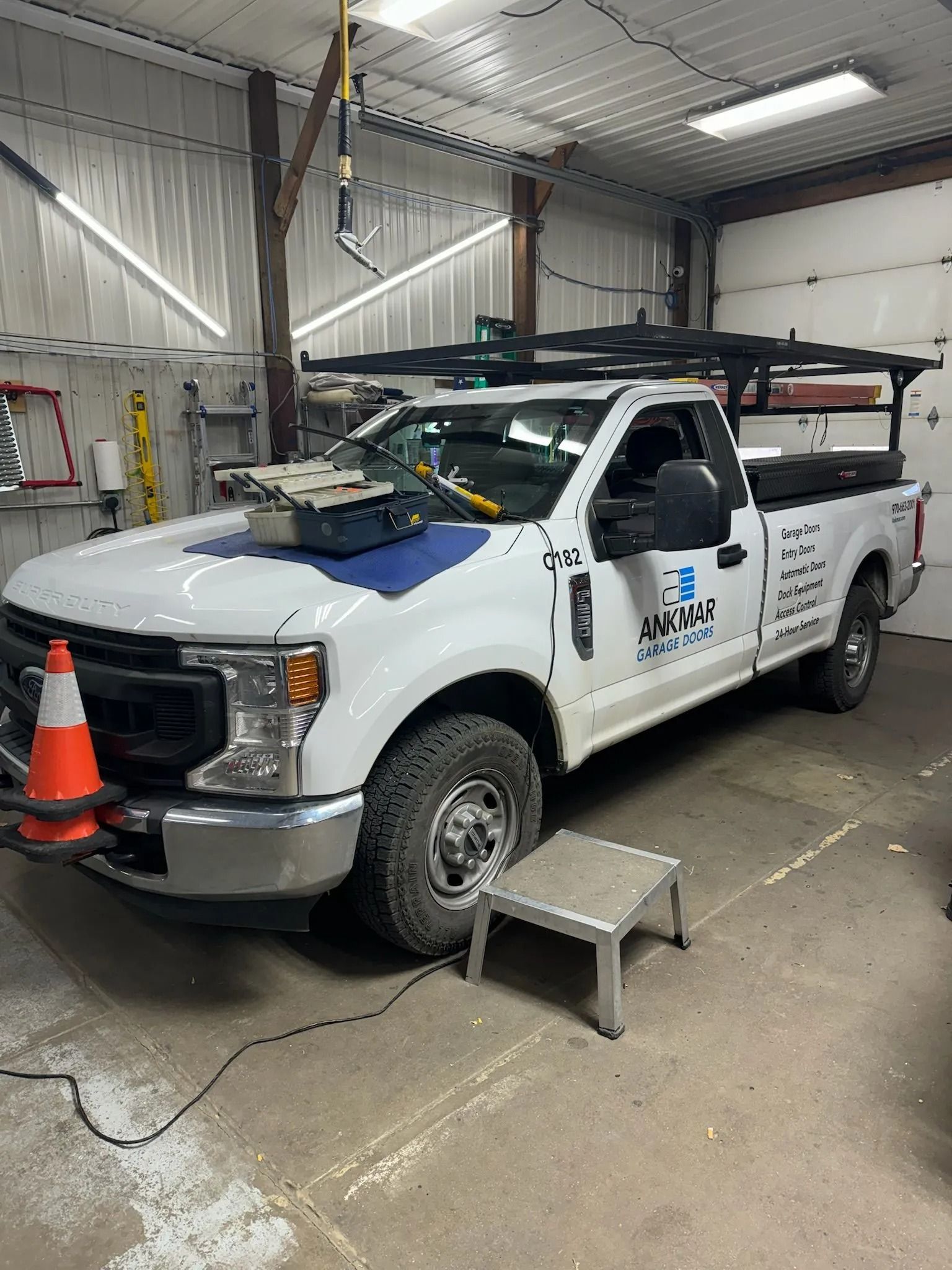 White work truck with a rack, cone, and step stool inside a garage.