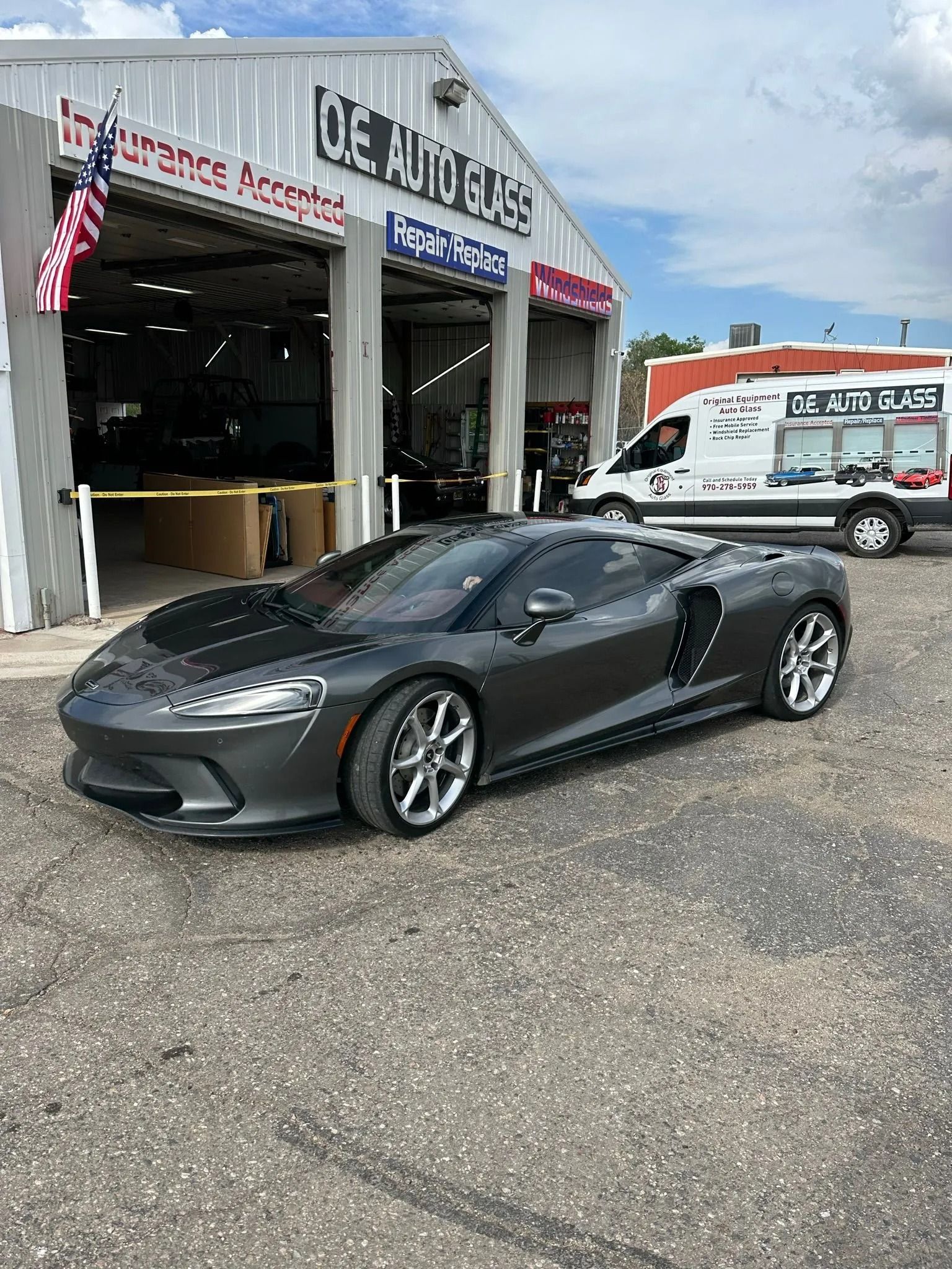 Gray sports car parked in front of an auto shop, with a white van in the background.