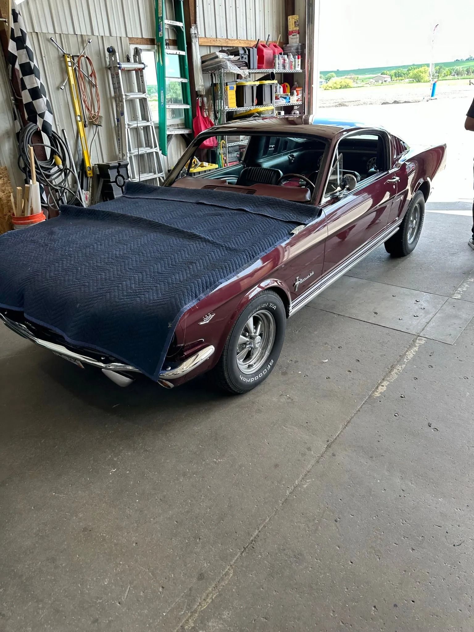 Burgundy classic Ford Mustang in a garage, covered hood. Shiny chrome accents, black and white checkered flag visible.