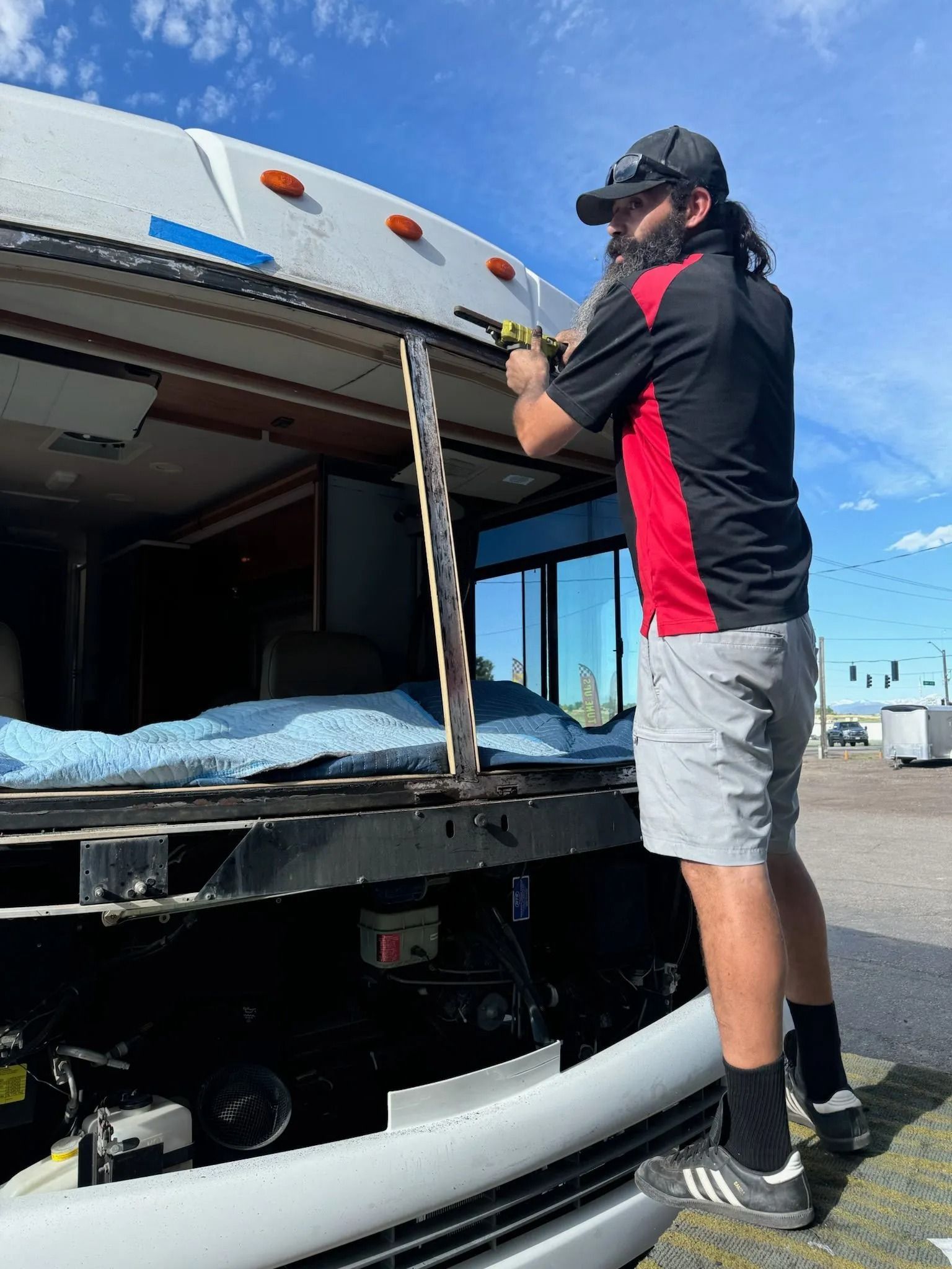 Man on a ladder repairs an RV's roof. He wears a hat, work shirt, and shorts. Outdoors on a sunny day.