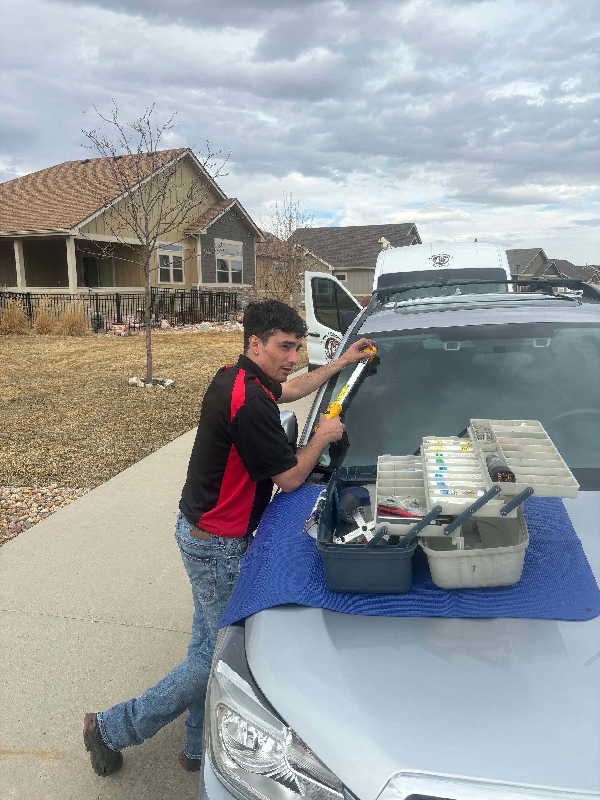Man replacing a windshield, using tools on a car in a residential area, under a cloudy sky.