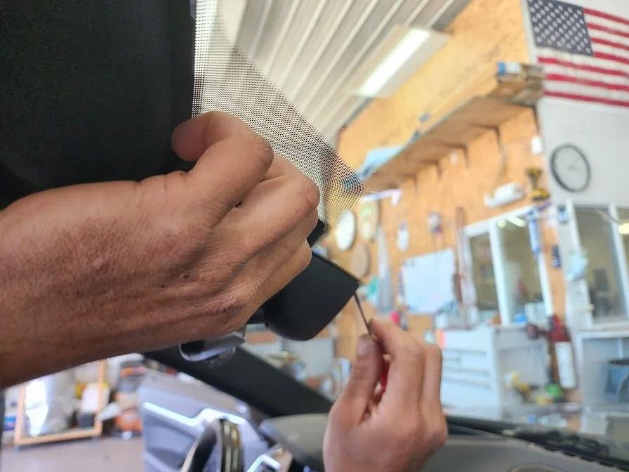 Hands installing a black device near a car's windshield. A small tool is used. An American flag is visible in the background.