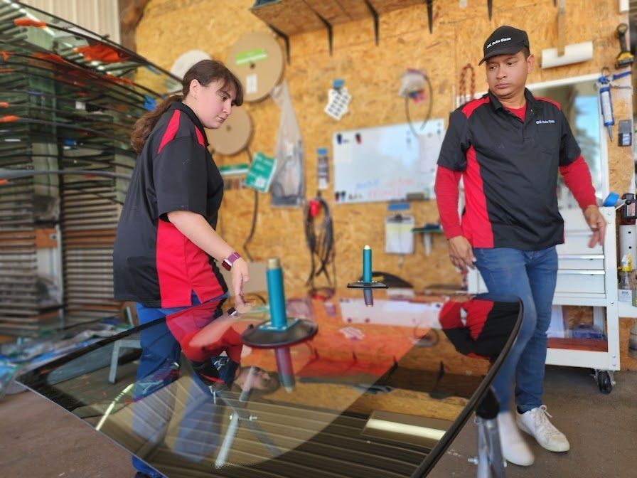 Two people in a shop setting installing a car windshield. They wear red and black shirts.