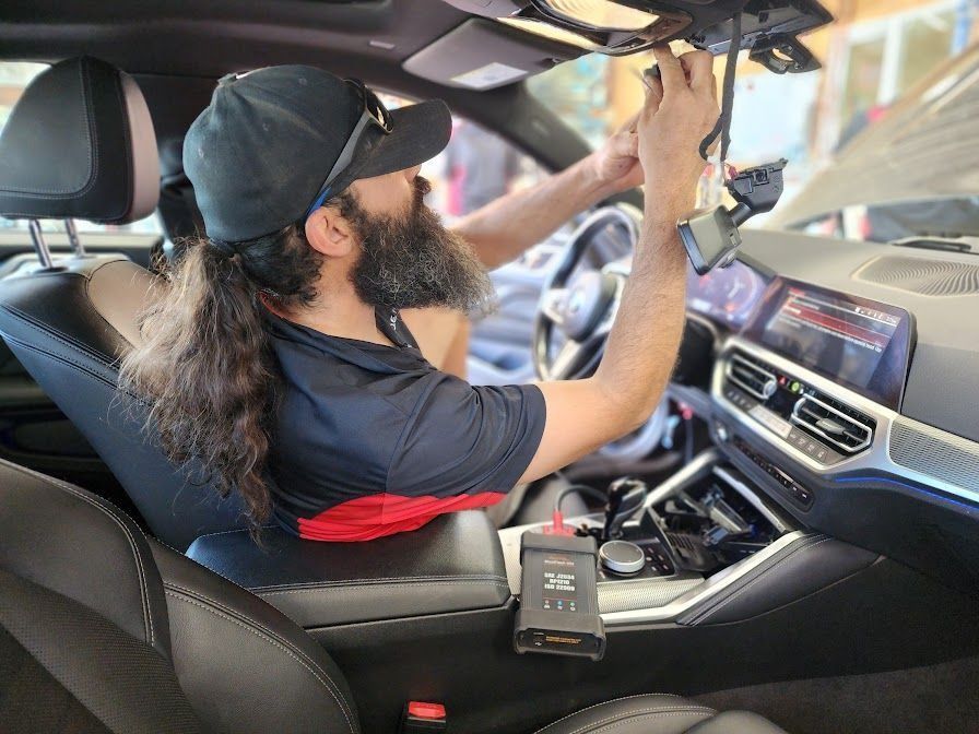 Man working on car interior, installing electronics. Black cap, grey beard, black leather seats.