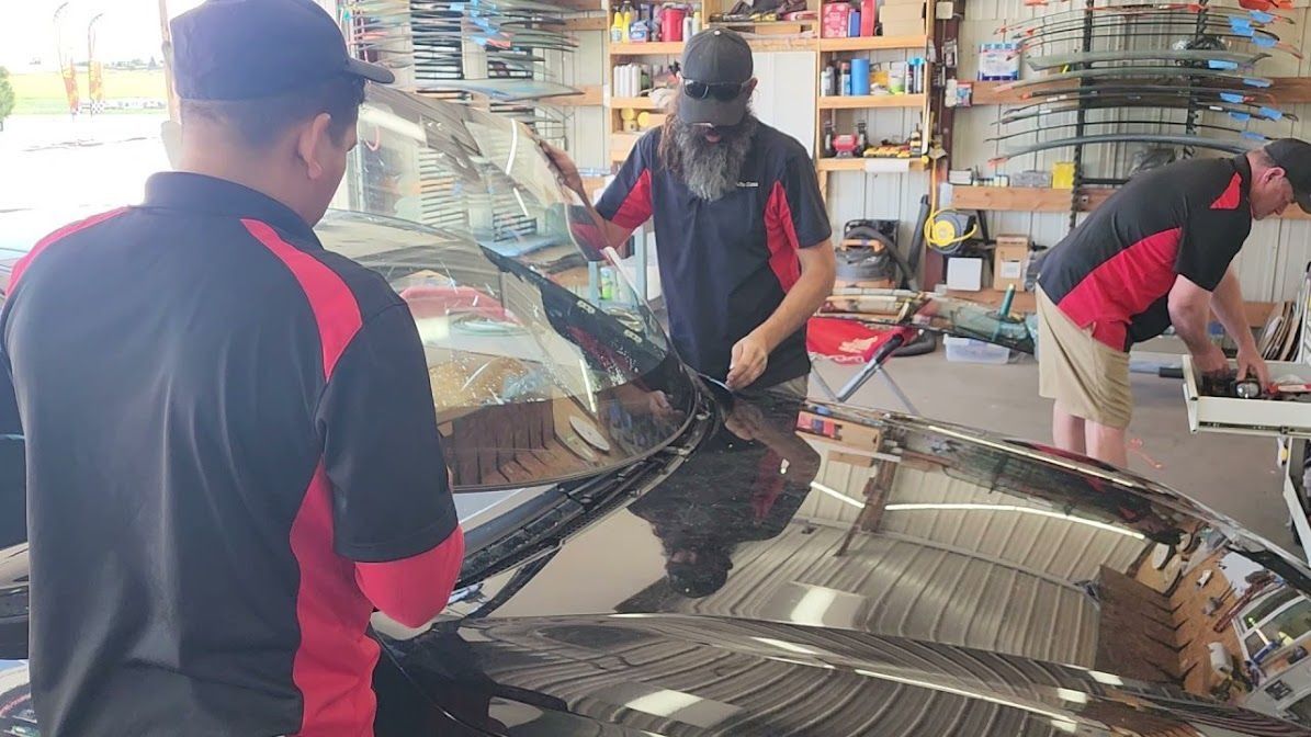 Three people installing window tint in a garage; black car, black and red shirts, hats, tools.