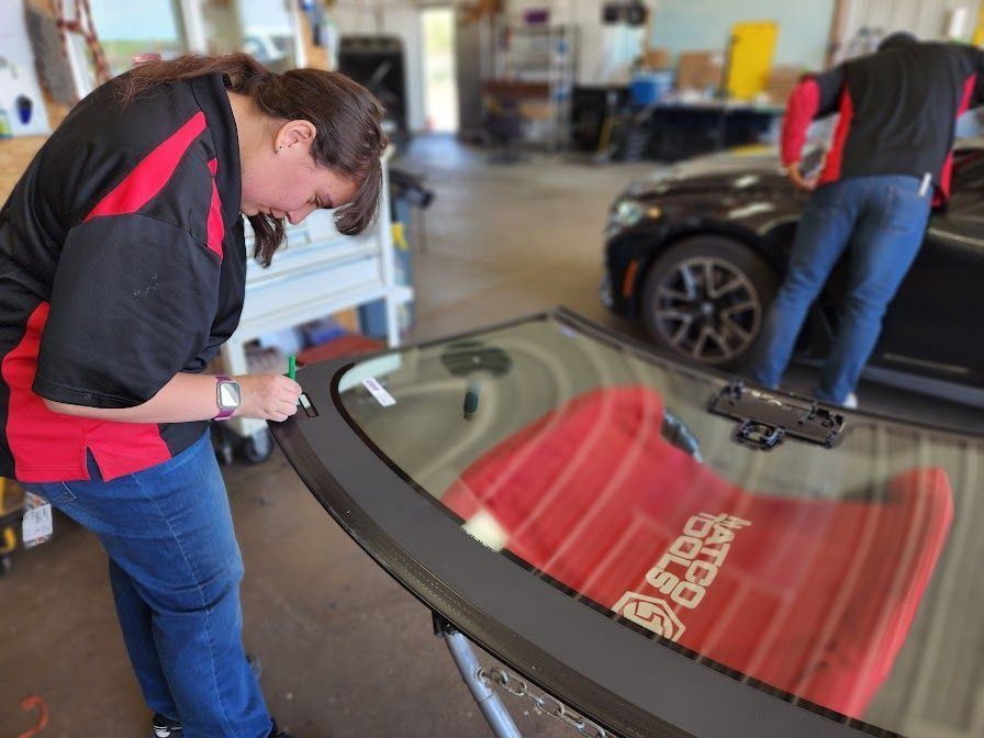 A person working on a windshield in a garage. Another person working on a black car in background.