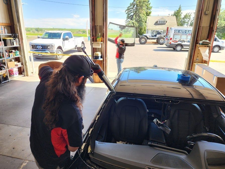 Workers replacing a car windshield in a garage. One uses a tool, another raises a new windshield.