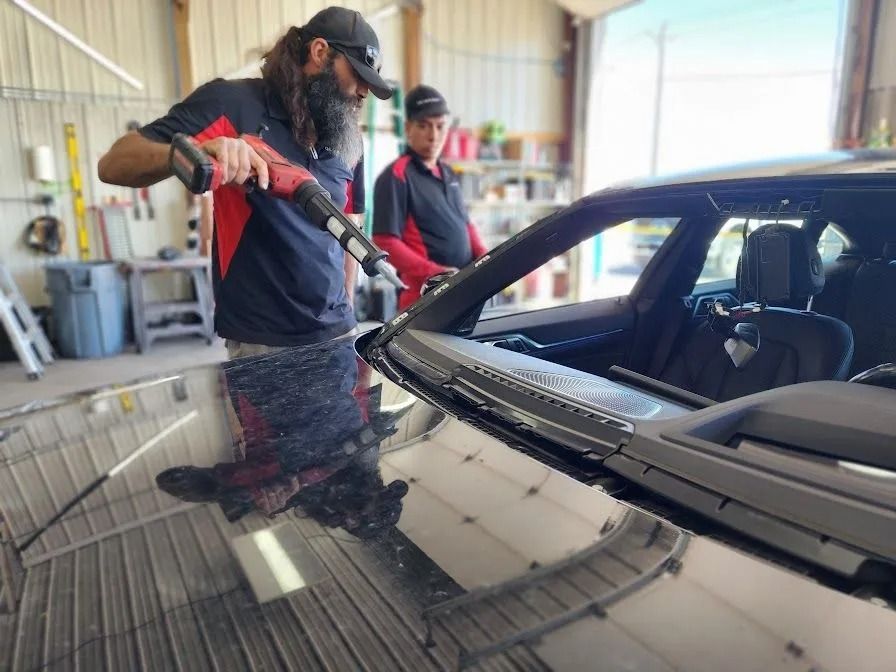 Two technicians replacing a car windshield inside a garage. One uses a tool to apply sealant.
