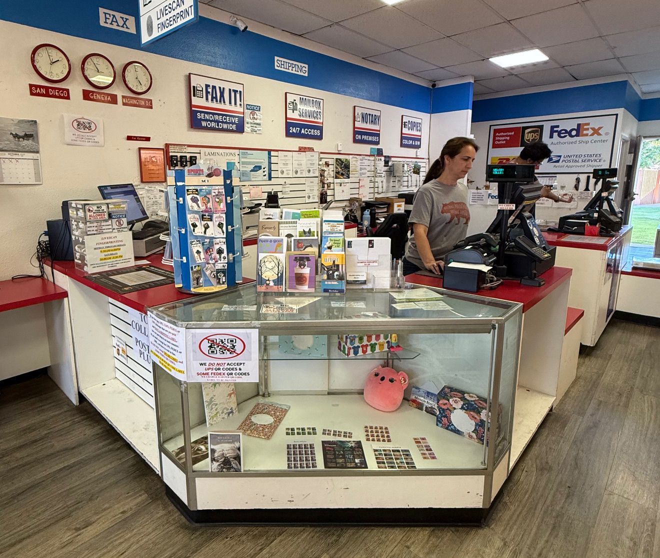 Inside a shop, a person behind the counter assists a customer. A display case is in front.