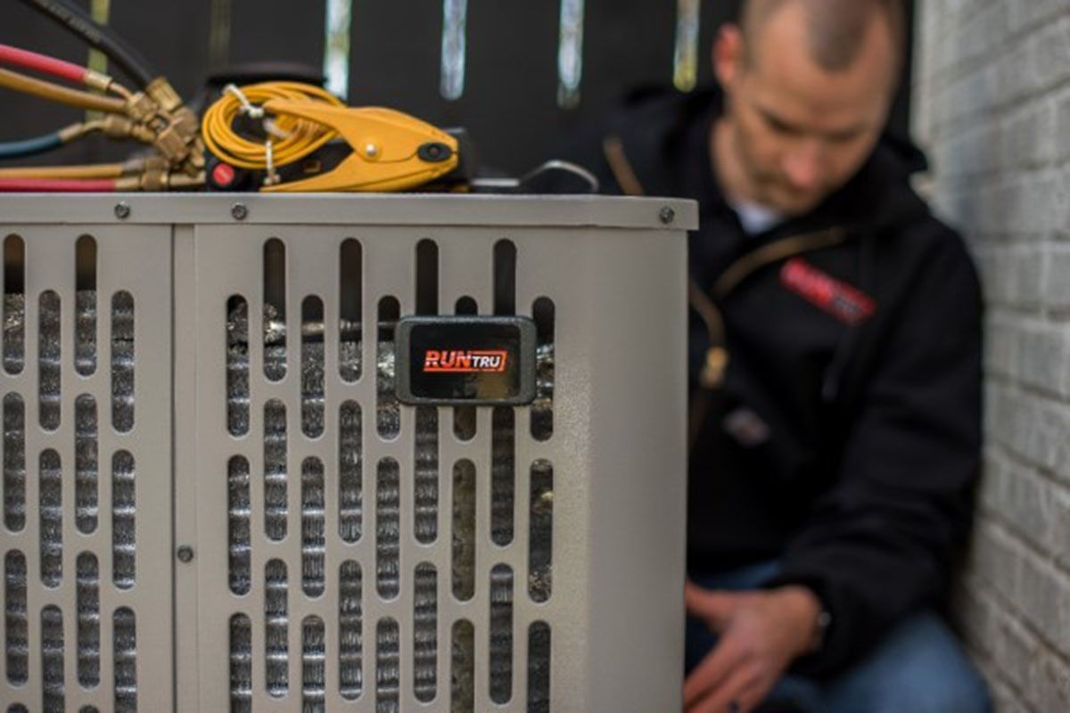 HVAC technician working on an outdoor air conditioning unit. A red-screened device is attached.