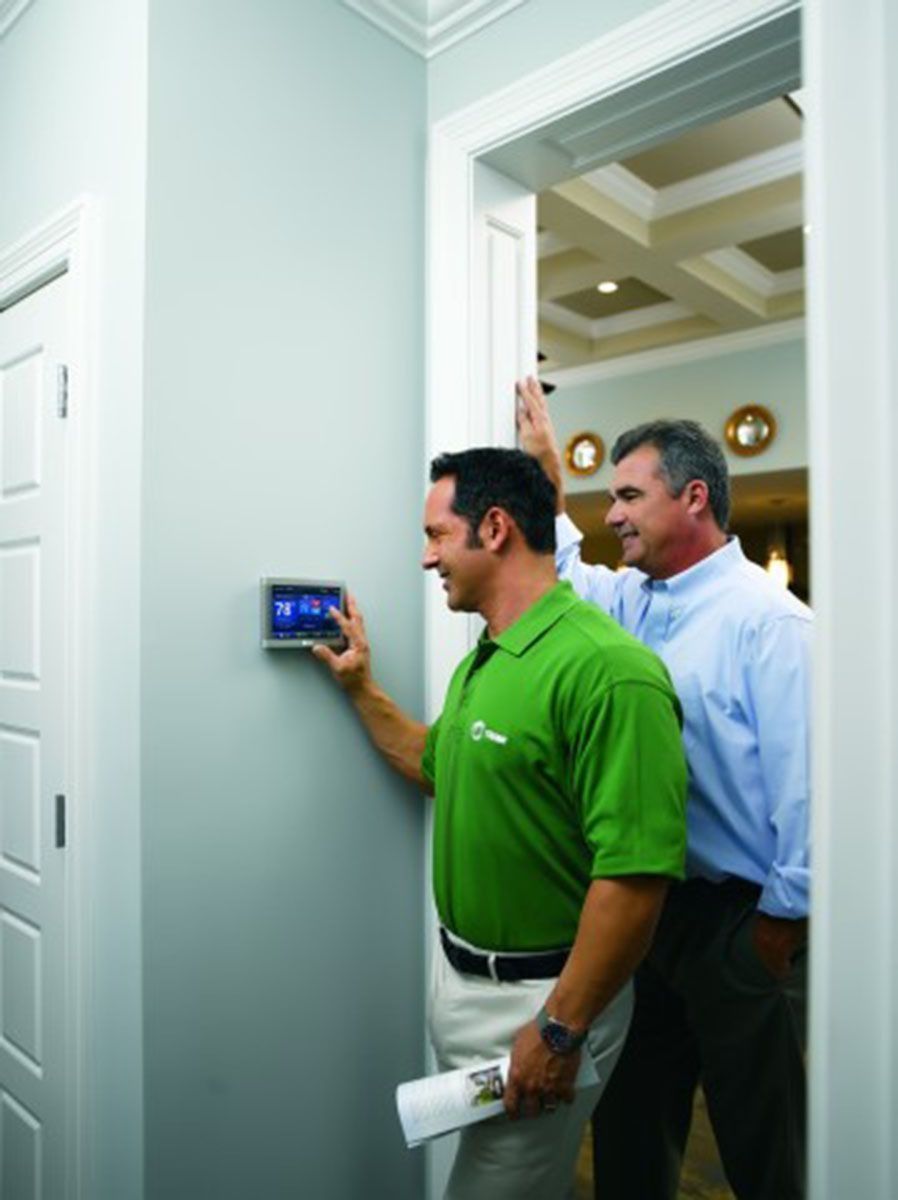 Two men looking at a thermostat on a wall. One man points, other smiles.