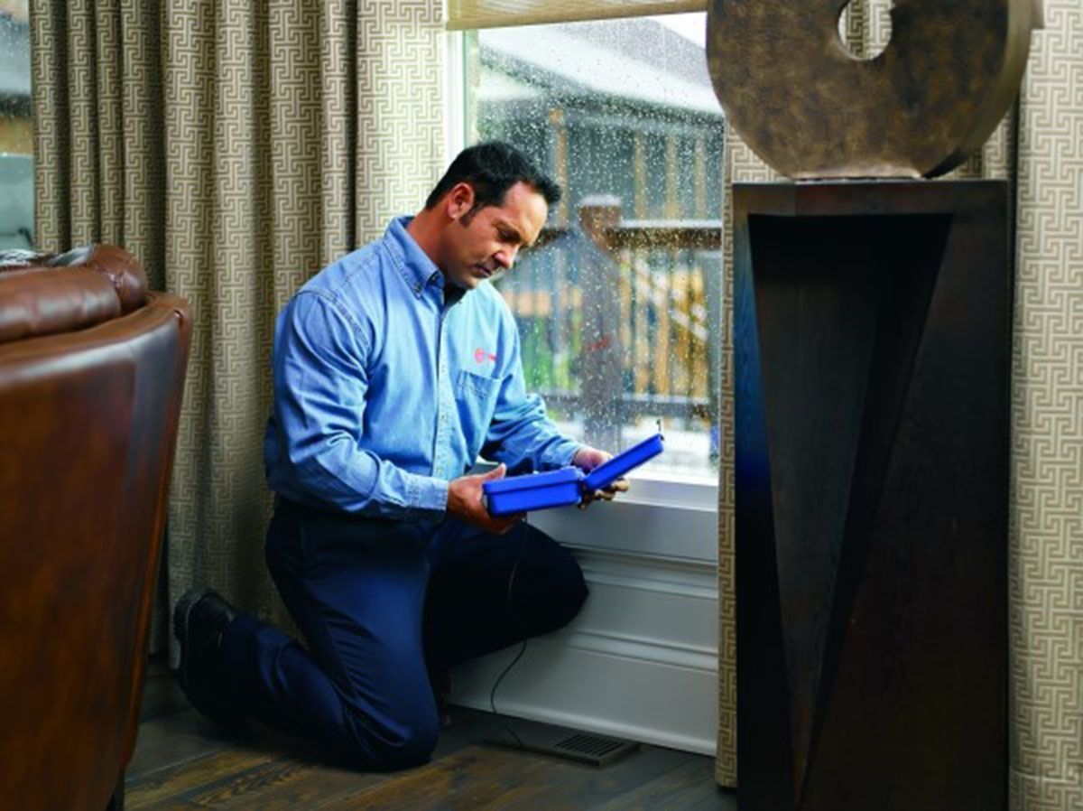 Man in blue uniform kneeling, examining a window. Inside a house, holding a blue box, looking down.