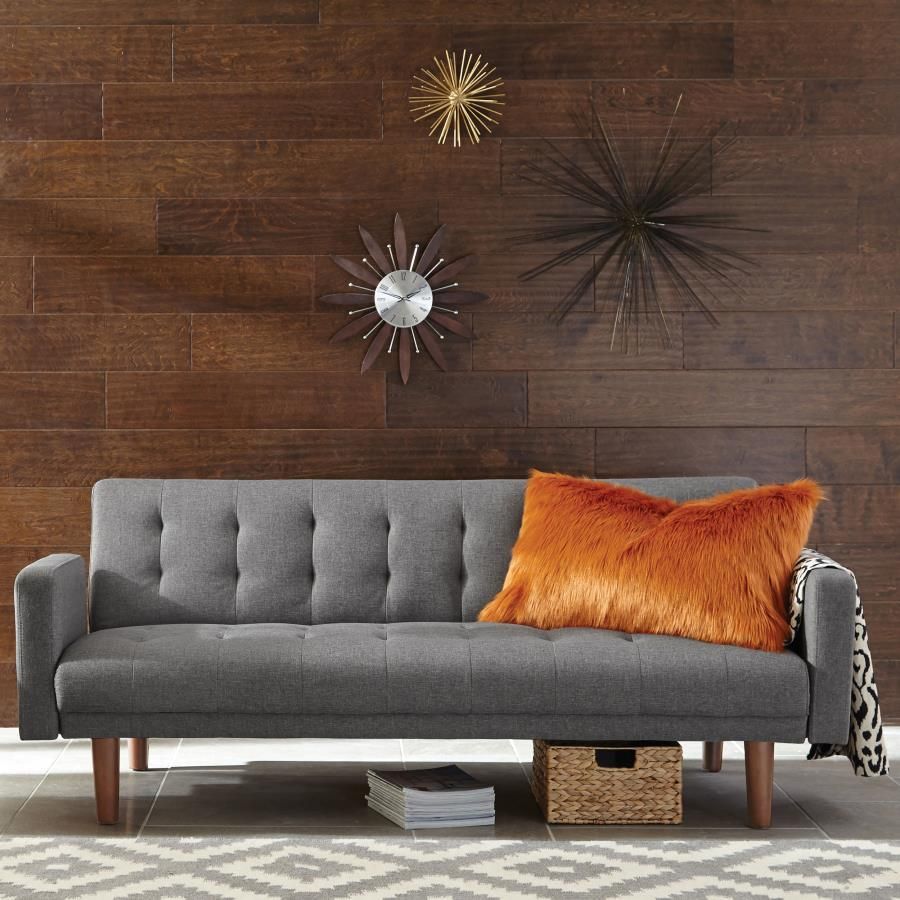 Gray futon sofa with orange pillow and wall decor against a wood paneled wall.