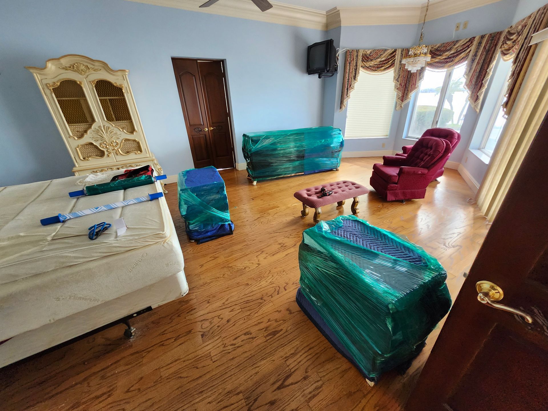 A room with furniture wrapped in green plastic, including a bed, cabinet, and chairs, likely in preparation for a move.