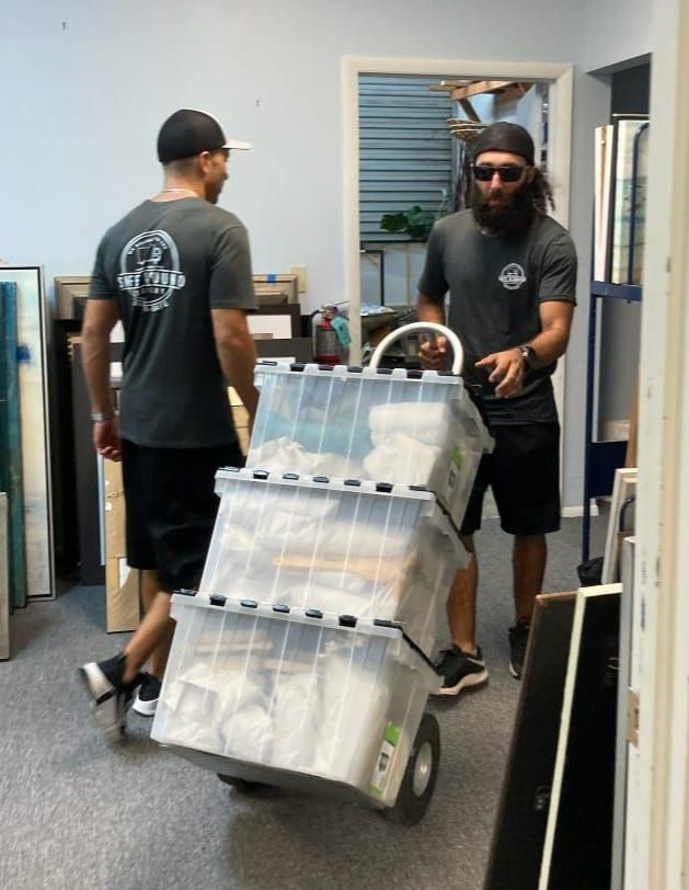 Two men move containers stacked on a hand truck in a room, one looks on.