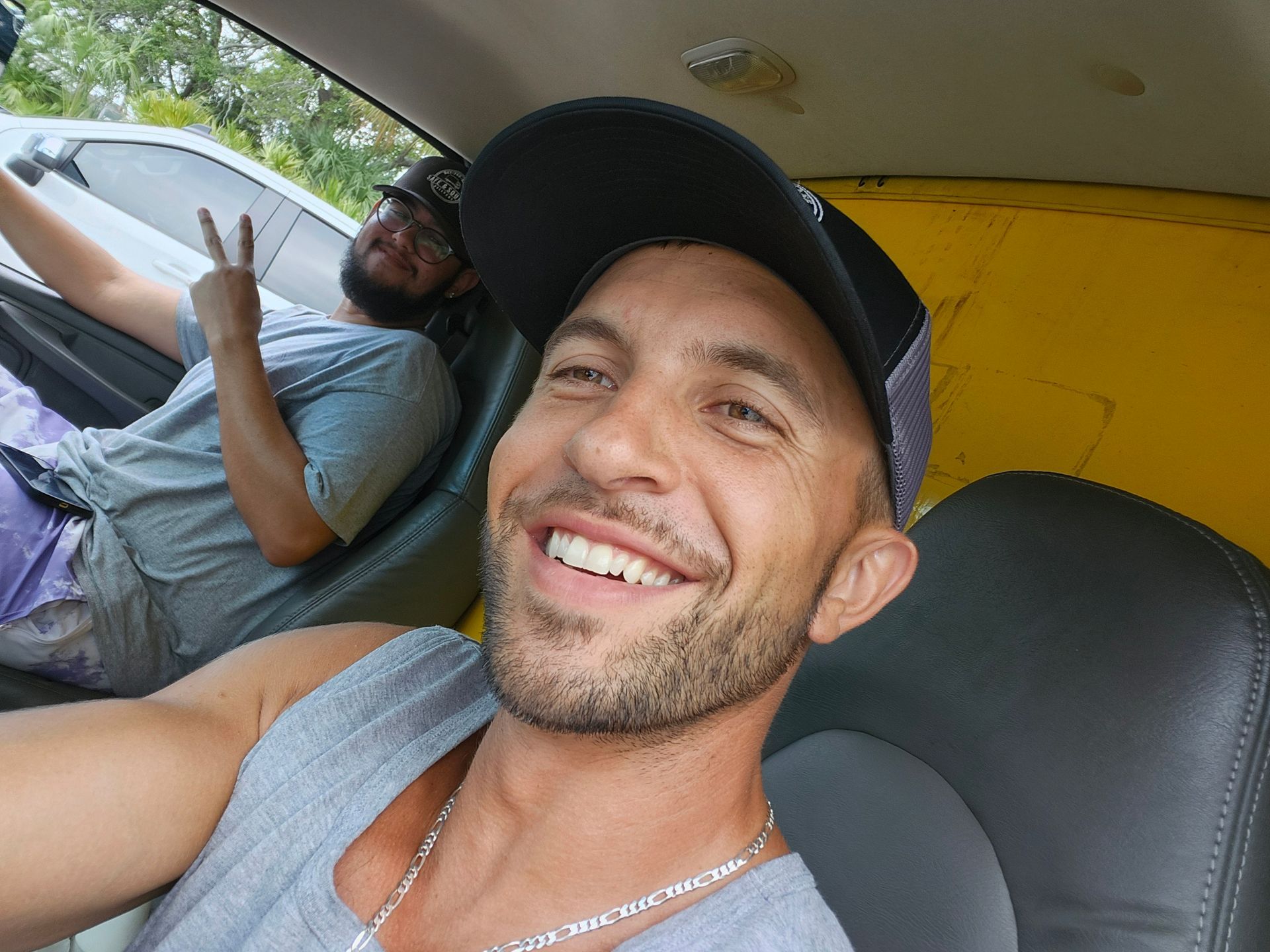 Two men in a yellow car, one smiling at the camera, the other making a peace sign.