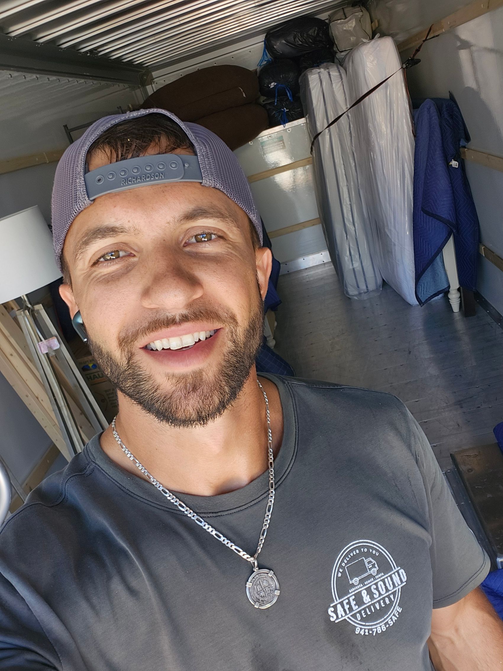 Smiling man in a cap inside a moving truck, surrounded by furniture and boxes.