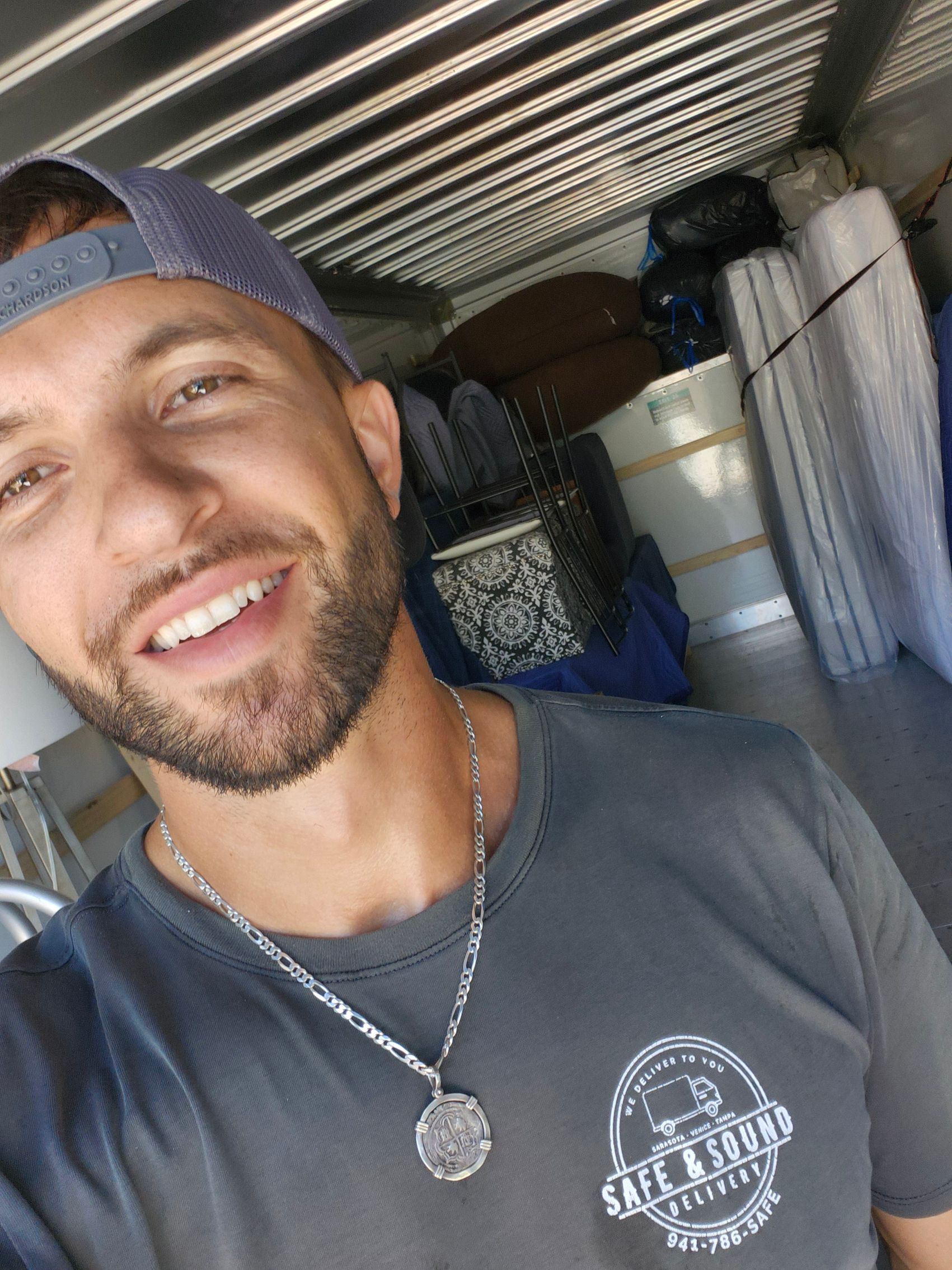 Man in gray shirt smiles, with cap in a moving truck filled with furniture.