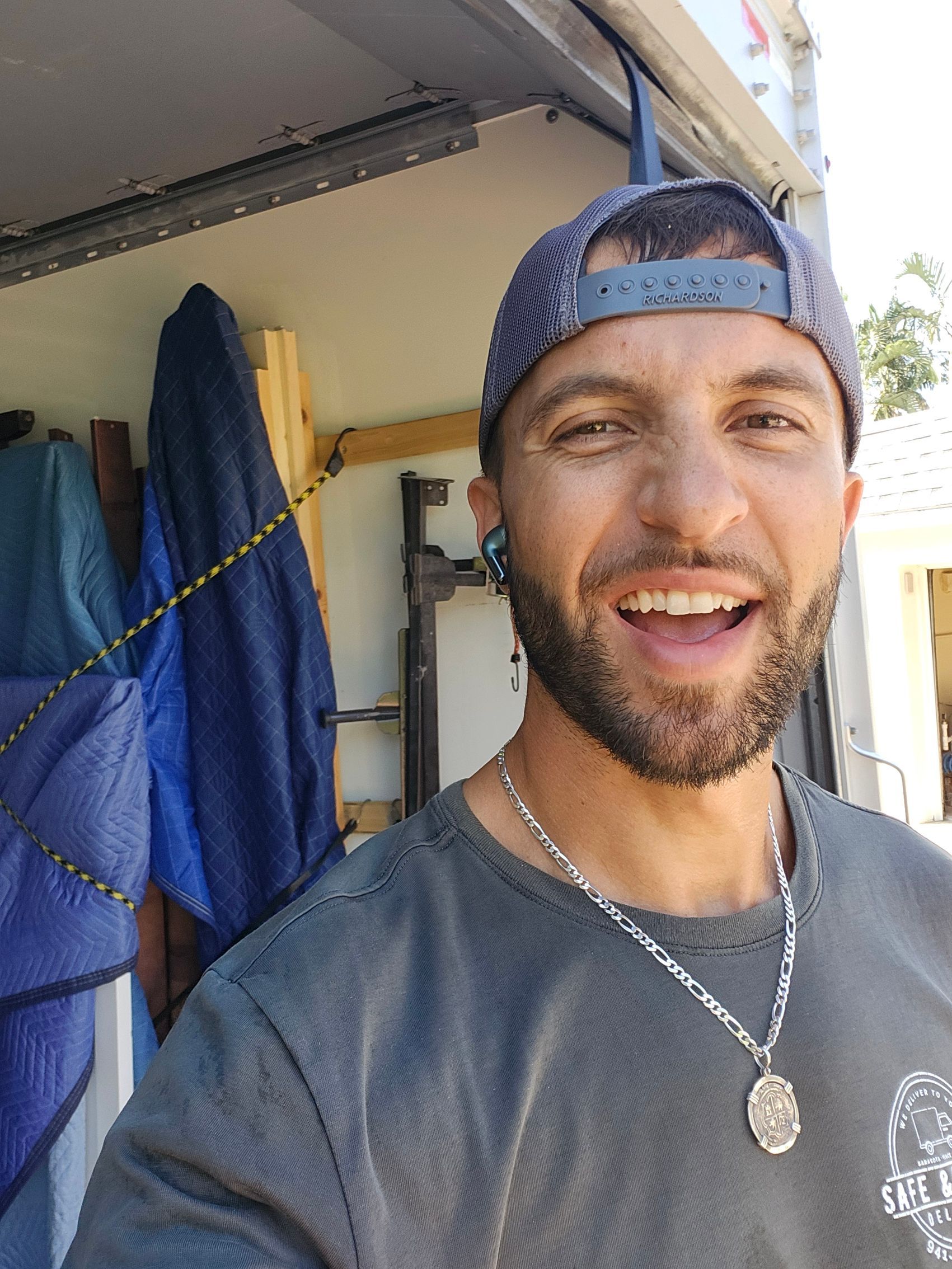Man smiling, wearing a cap, gray shirt, and silver necklace in a garage.