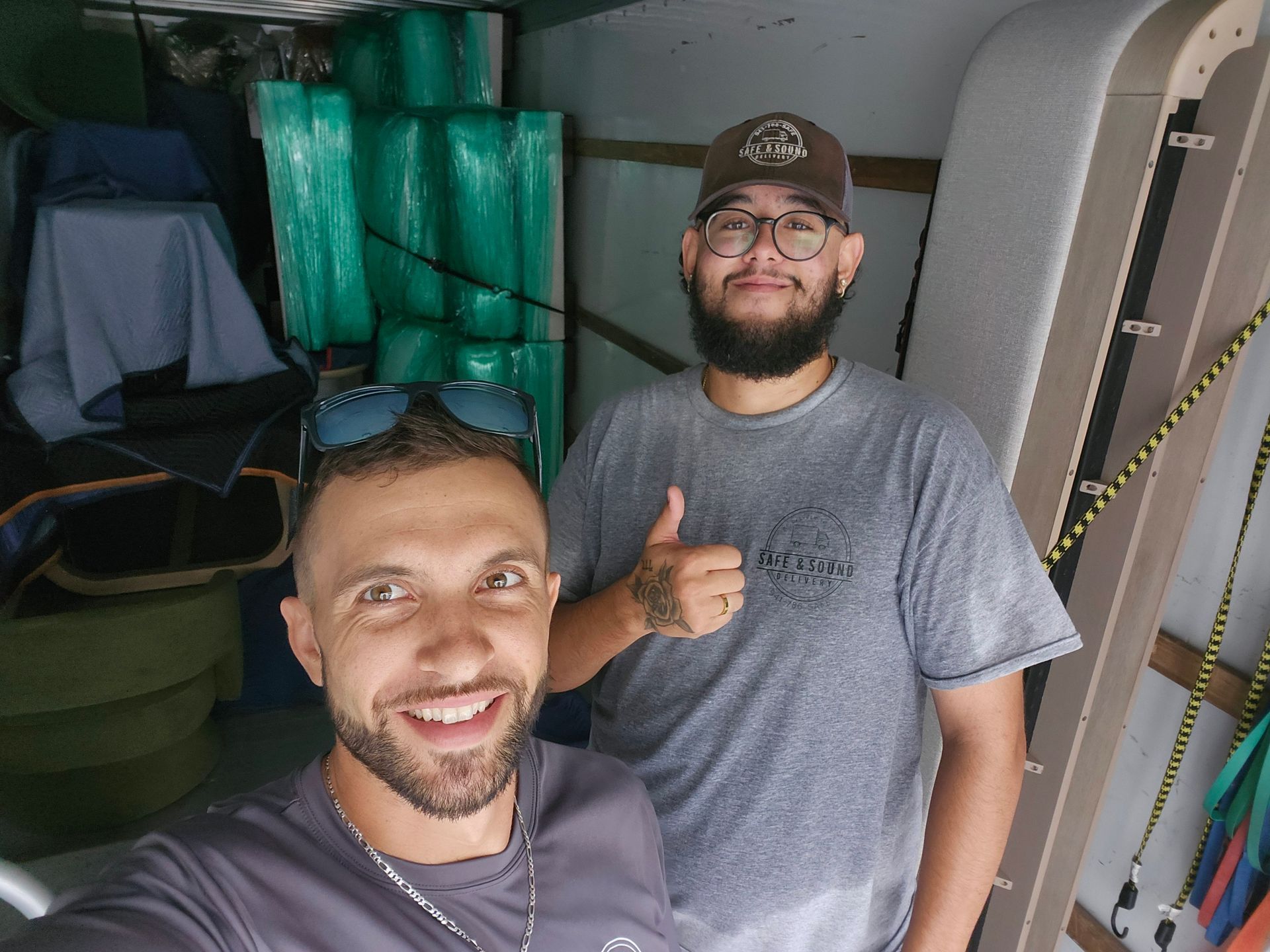 Two men smiling, one giving a thumbs-up, inside a truck with construction materials in the background.