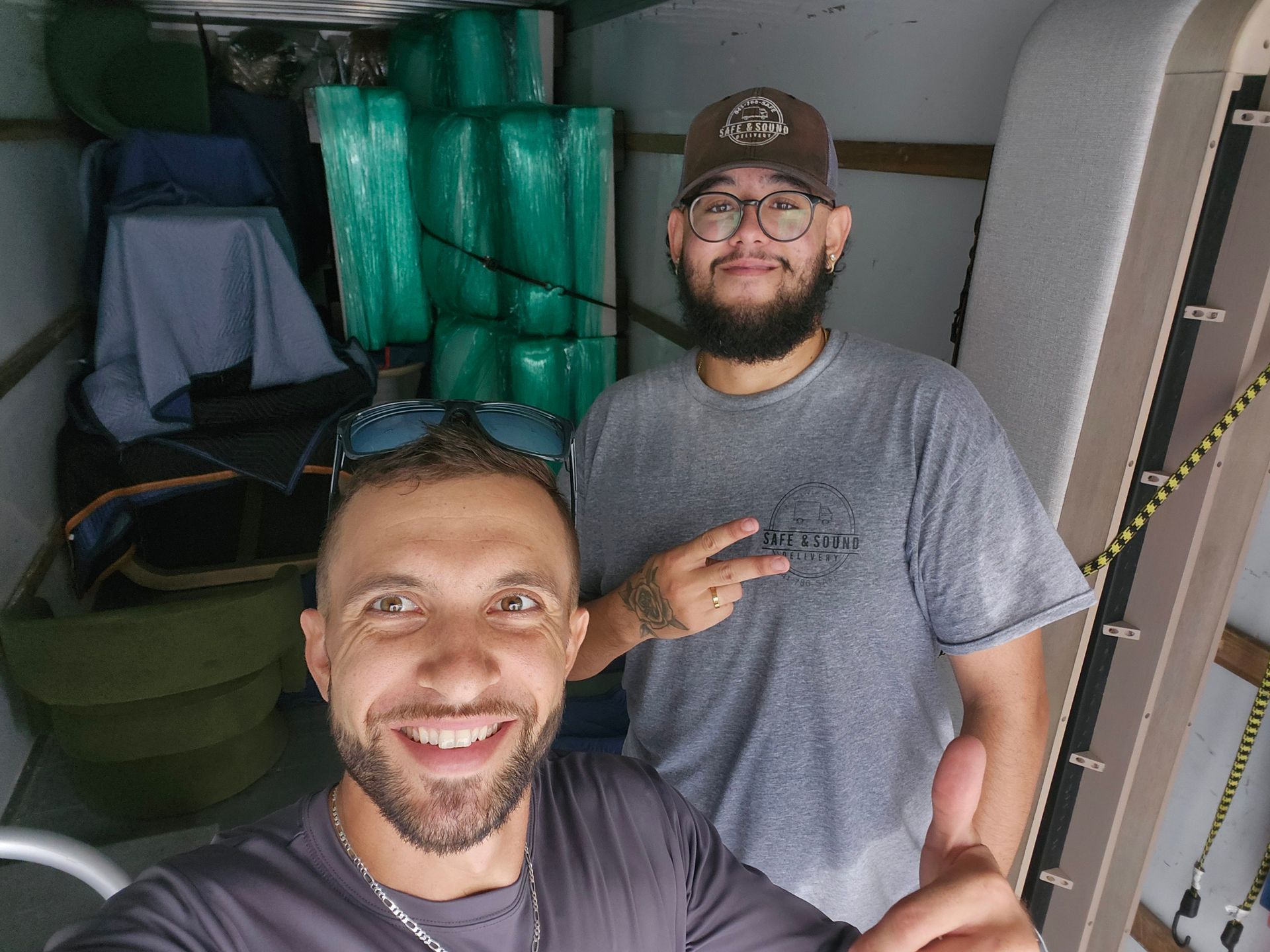 Two men smile in a moving truck full of items. One gives a thumbs up, the other a peace sign.