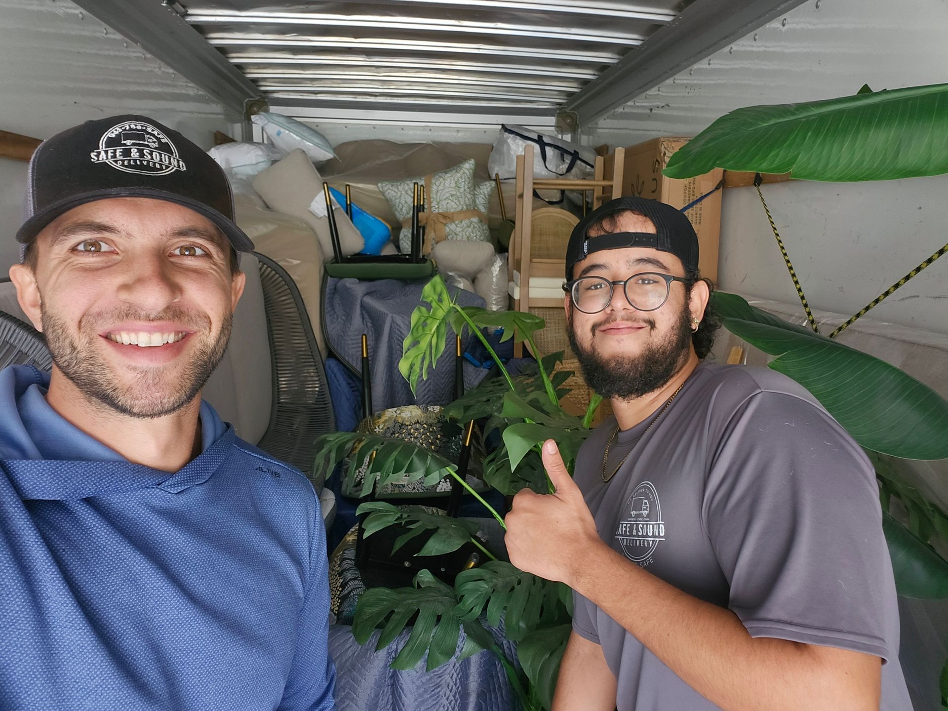 Two men smiling, inside a moving truck. One gives a thumbs up, holding a large plant.