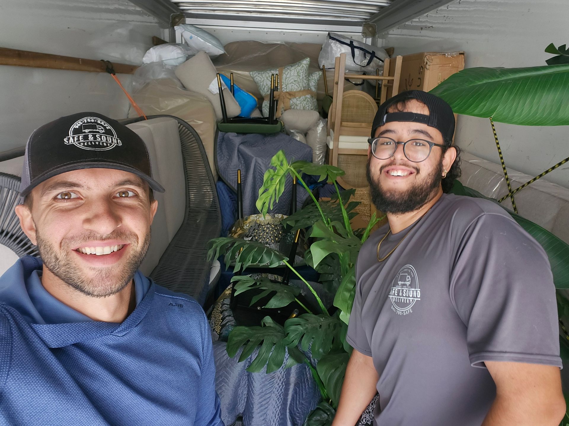 Two men smile inside a packed moving truck. Furniture and plants fill the cargo area.