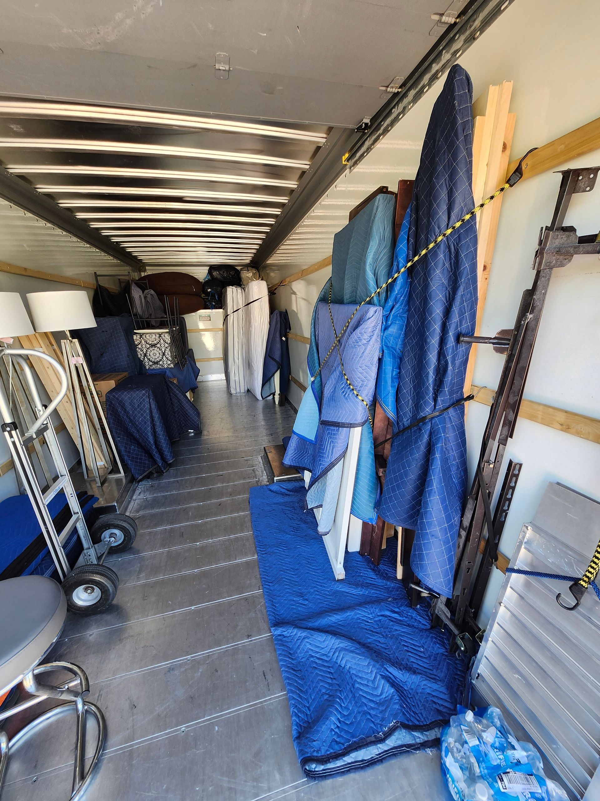 Inside a moving truck filled with packed blue and white furniture, lamps, and items for transport.