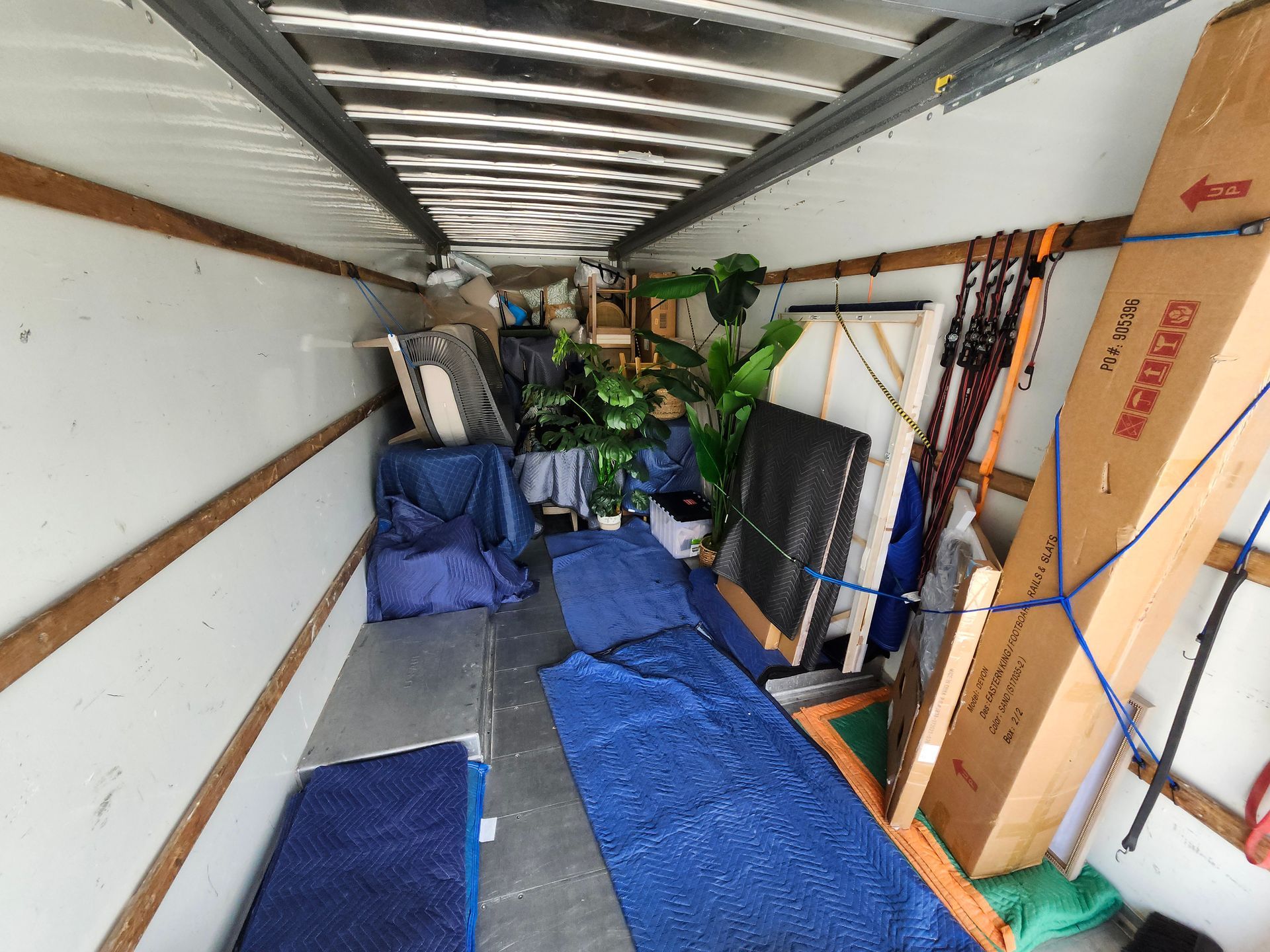 Interior of a moving truck filled with furniture, plants, and boxes. Blue pads cover the floor.