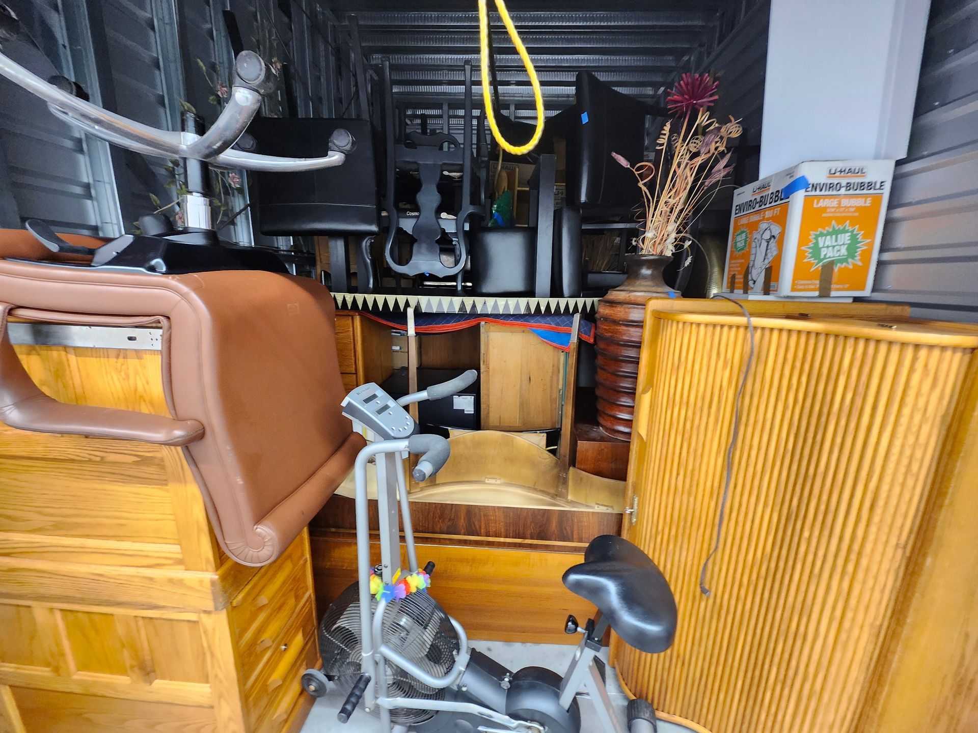 Storage unit filled with furniture, a desk, exercise bike, monitors, and boxes.