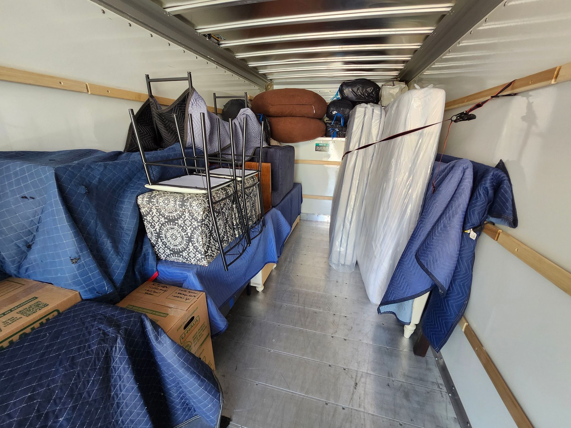 Inside of a moving truck packed with furniture, boxes, and a mattress, secured with blue moving blankets.