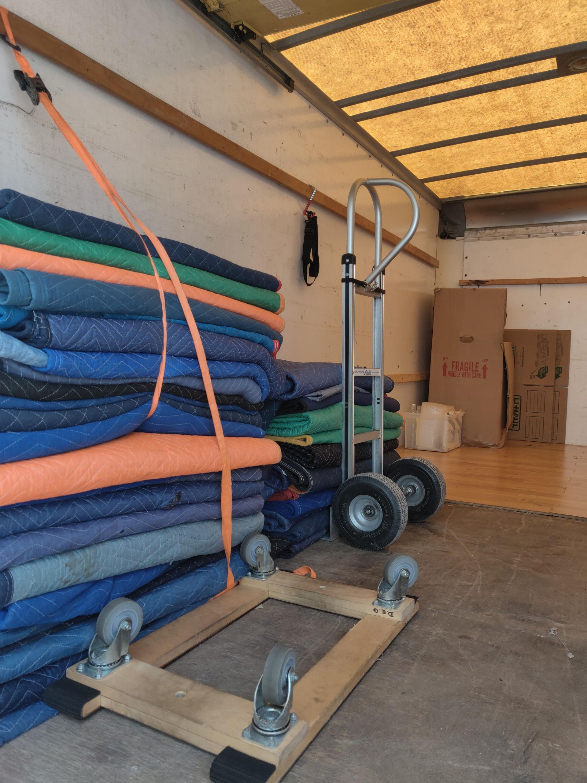 Inside a moving truck, a stack of blue moving blankets, a hand truck, and a wooden dolly.