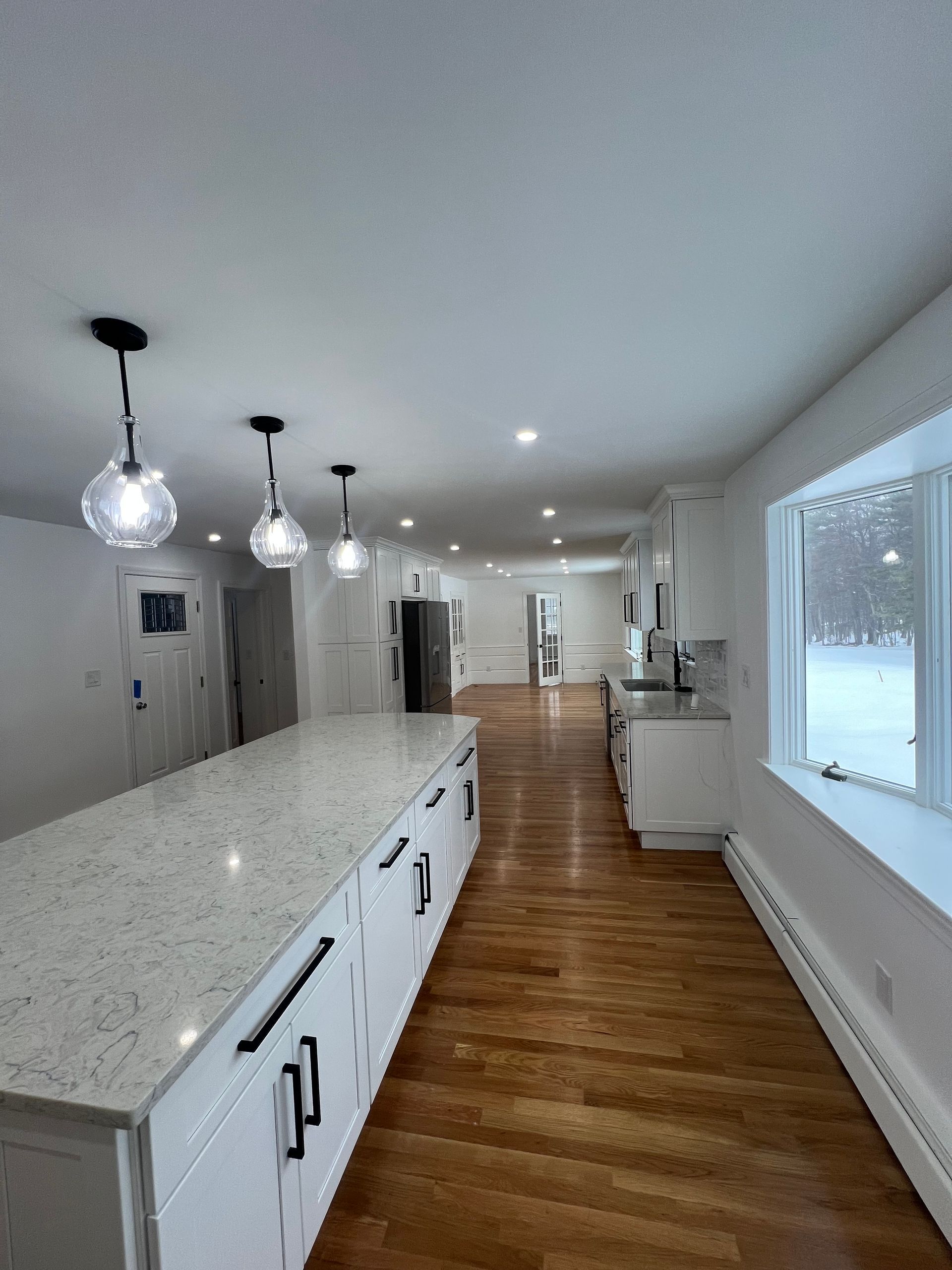 A bright, modern kitchen with a long white island, light granite countertops, hardwood floors, and pendant lighting.